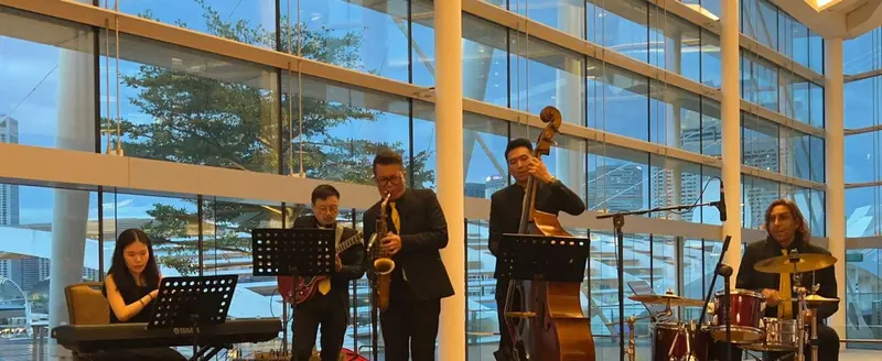 Five piece jazz band with keyboard, guitar, saxophone, double bass and drums playing in a glass atrium at dusk