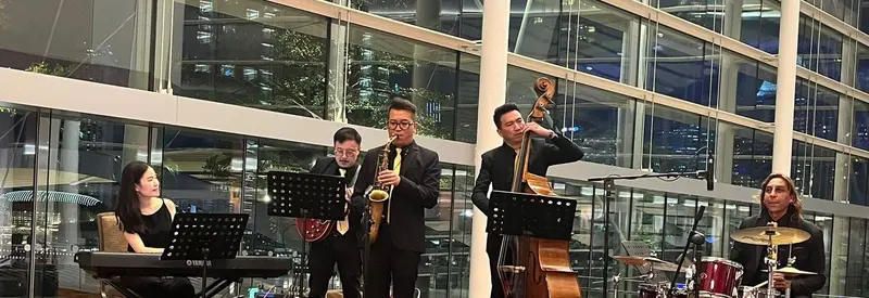 Jazz quintet with a saxophonist, guitarist, double bass player, keyboard player and drummer on a stage in a glass atrium at night